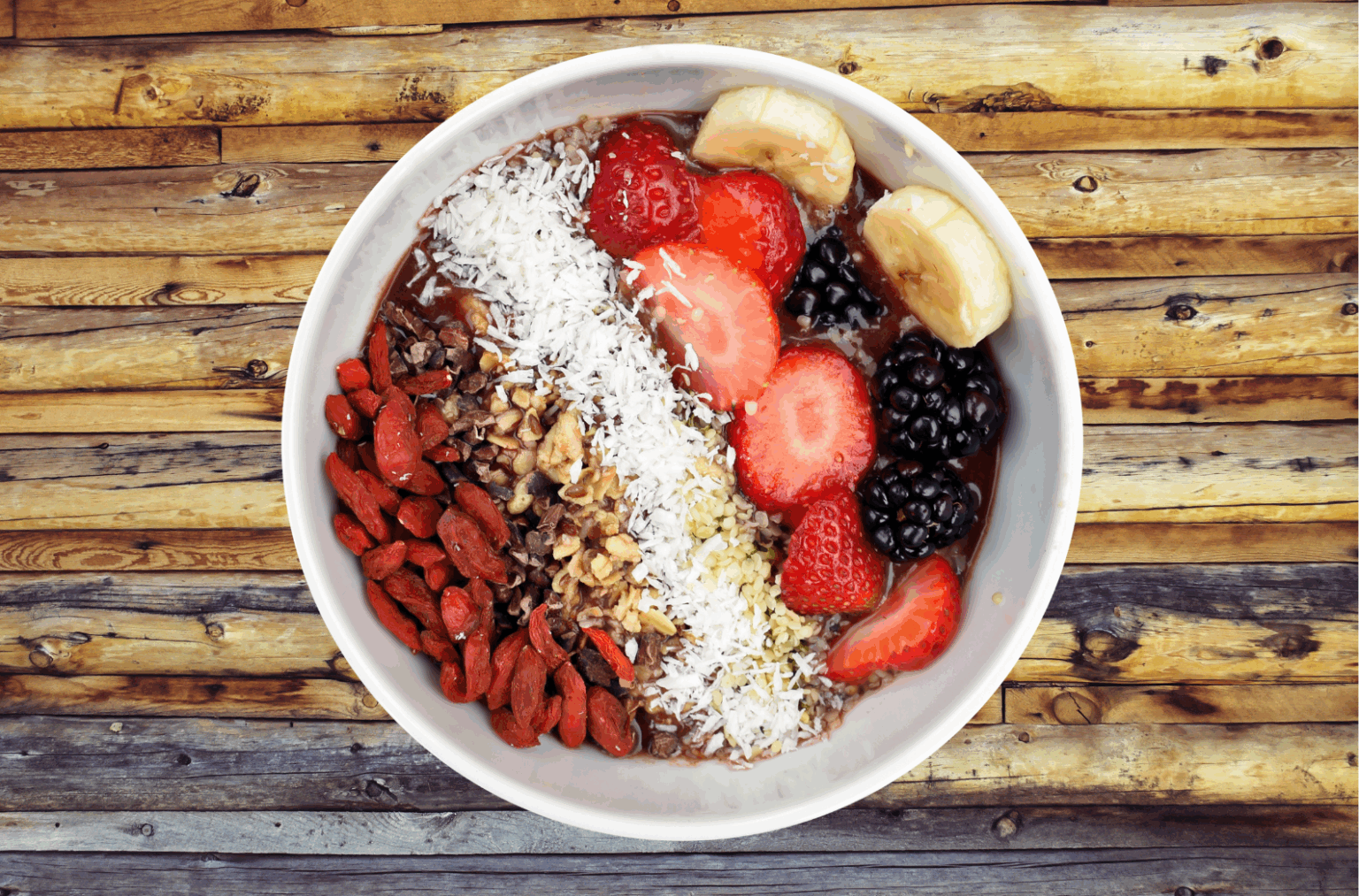 Bol coloré composé de fraises, bananes, mûres, baies de goji, noix, éclats de cacao et noix de coco râpée sur une base de smoothie, illustrant une collation saine et rassasiante idéale pour le cycle menstruel.